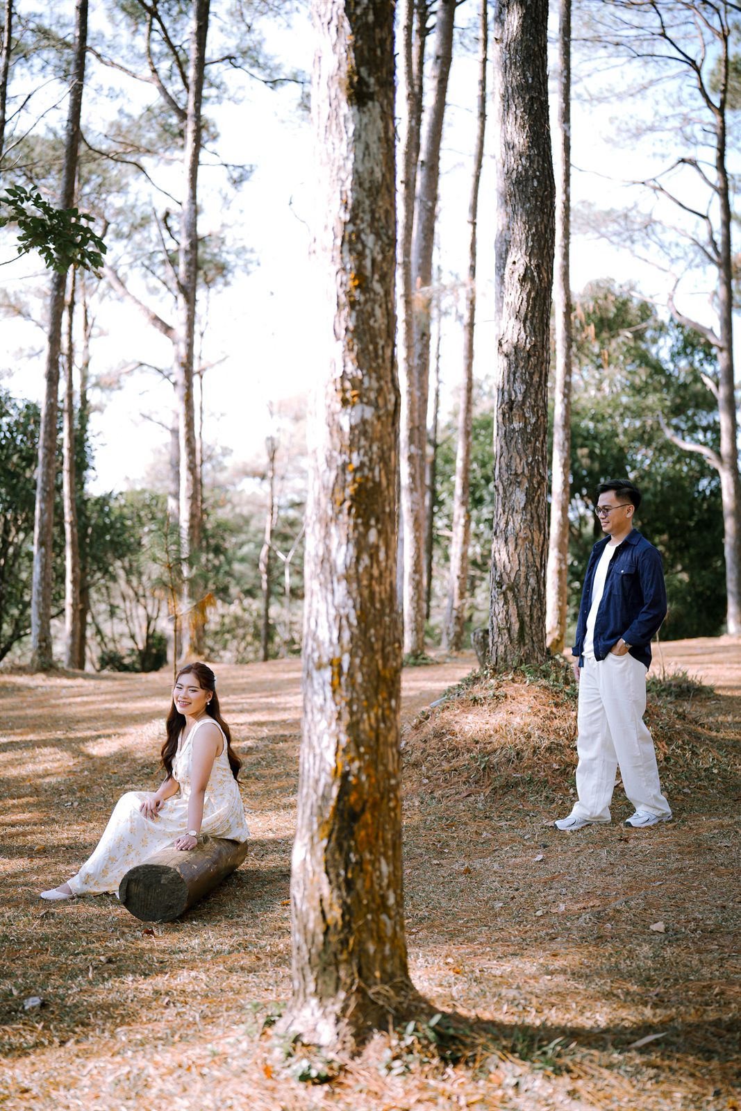 Caren seated on a log while Zayrol stands nearby in the pines